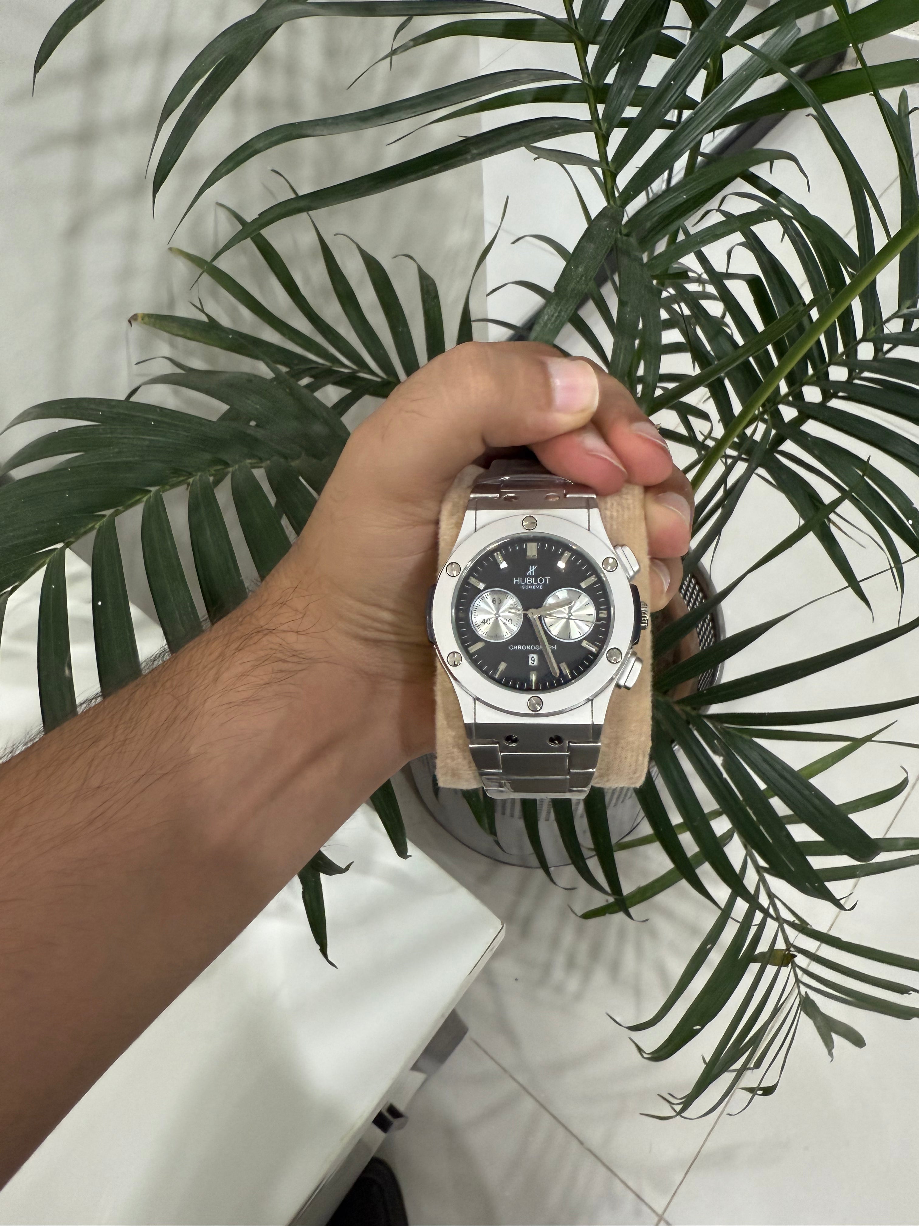 Hand holding a silver hublot watch with a black face against a leafy green background