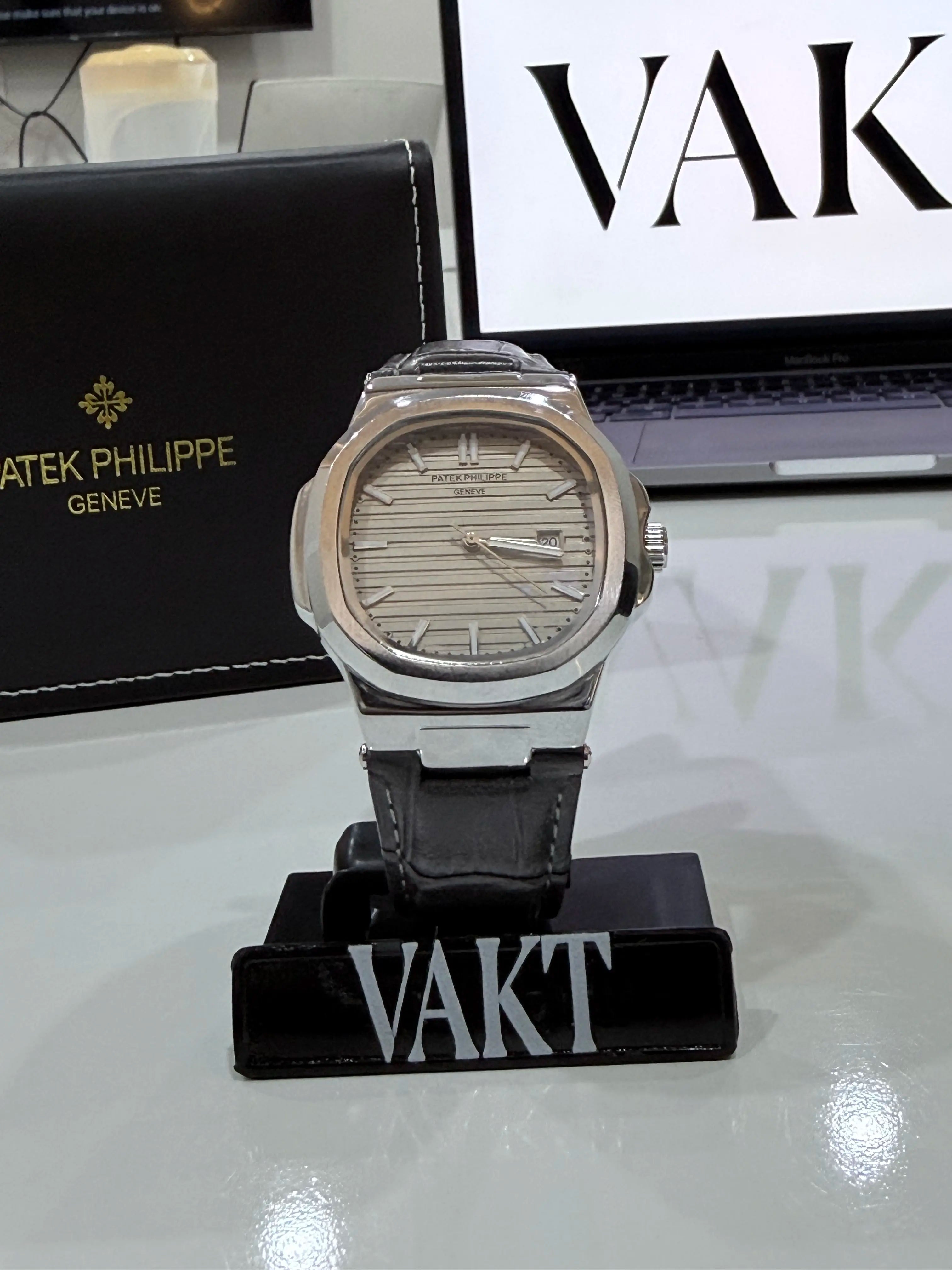 Watch on a stand with 'VAKT' branding, placed on a reflective surface with 'VAKT' and 'Patek Philippe' visible in the background.