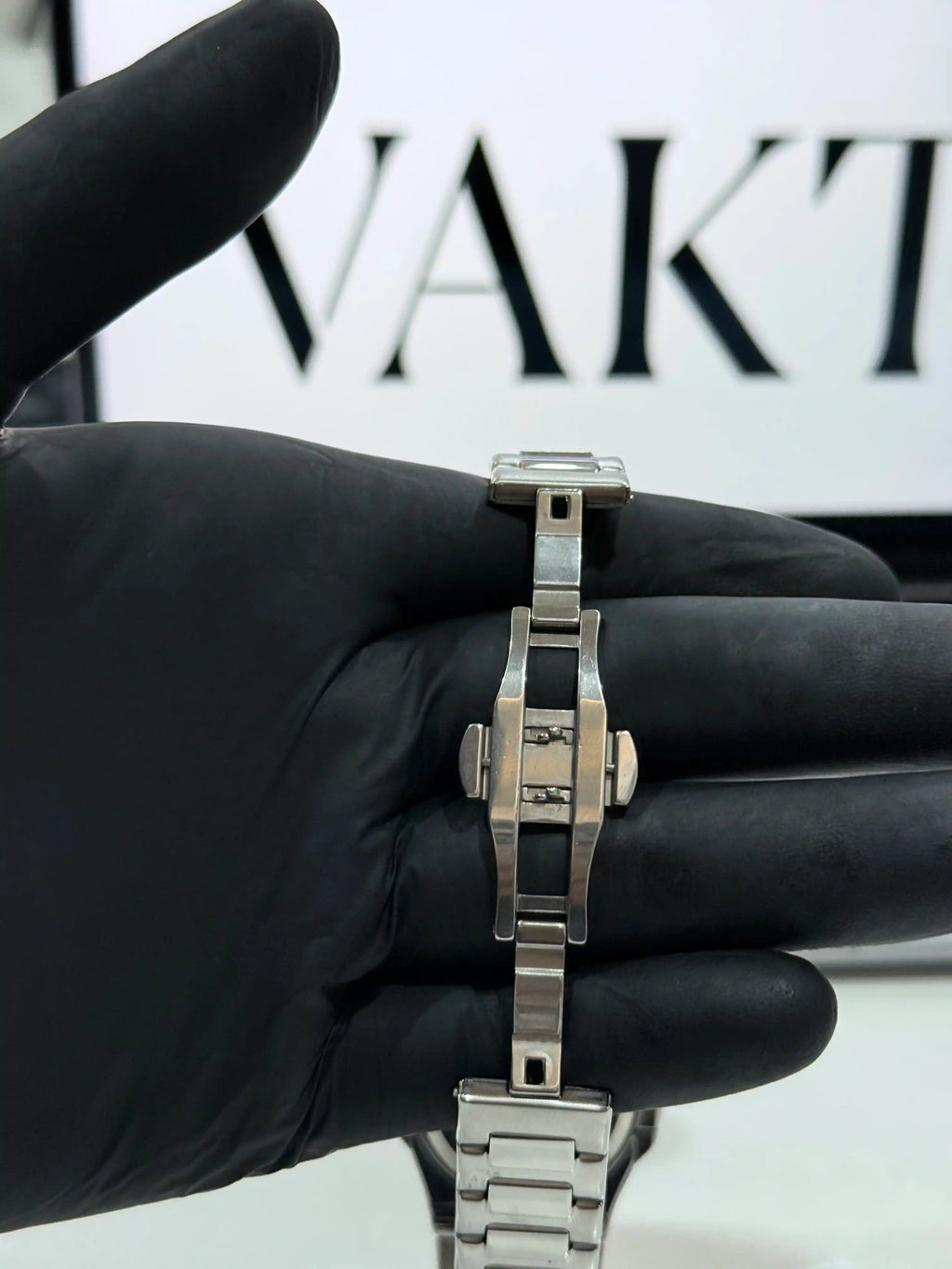 Silver watch on a hand wearing a black glove with 'vakt' branding in the background.