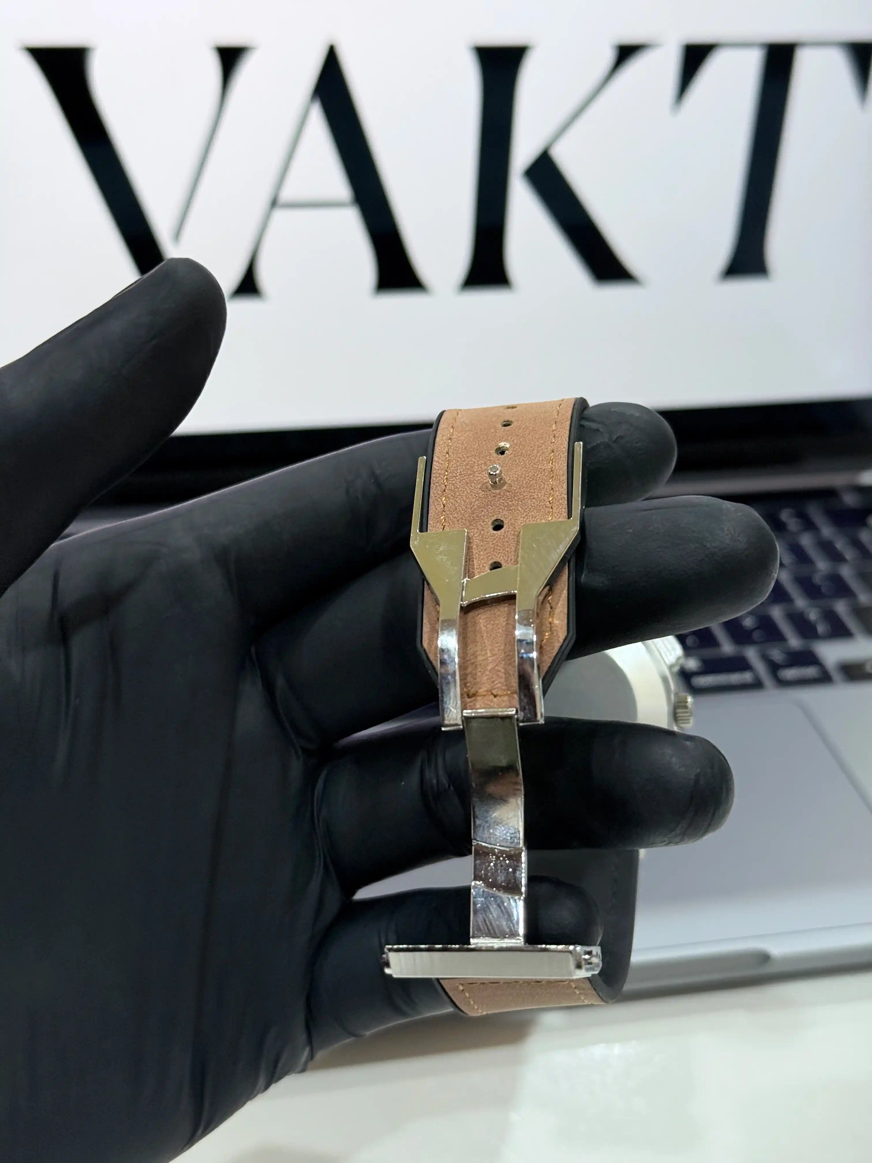Hand holding a watch strap with 'VAKT' branding in the background