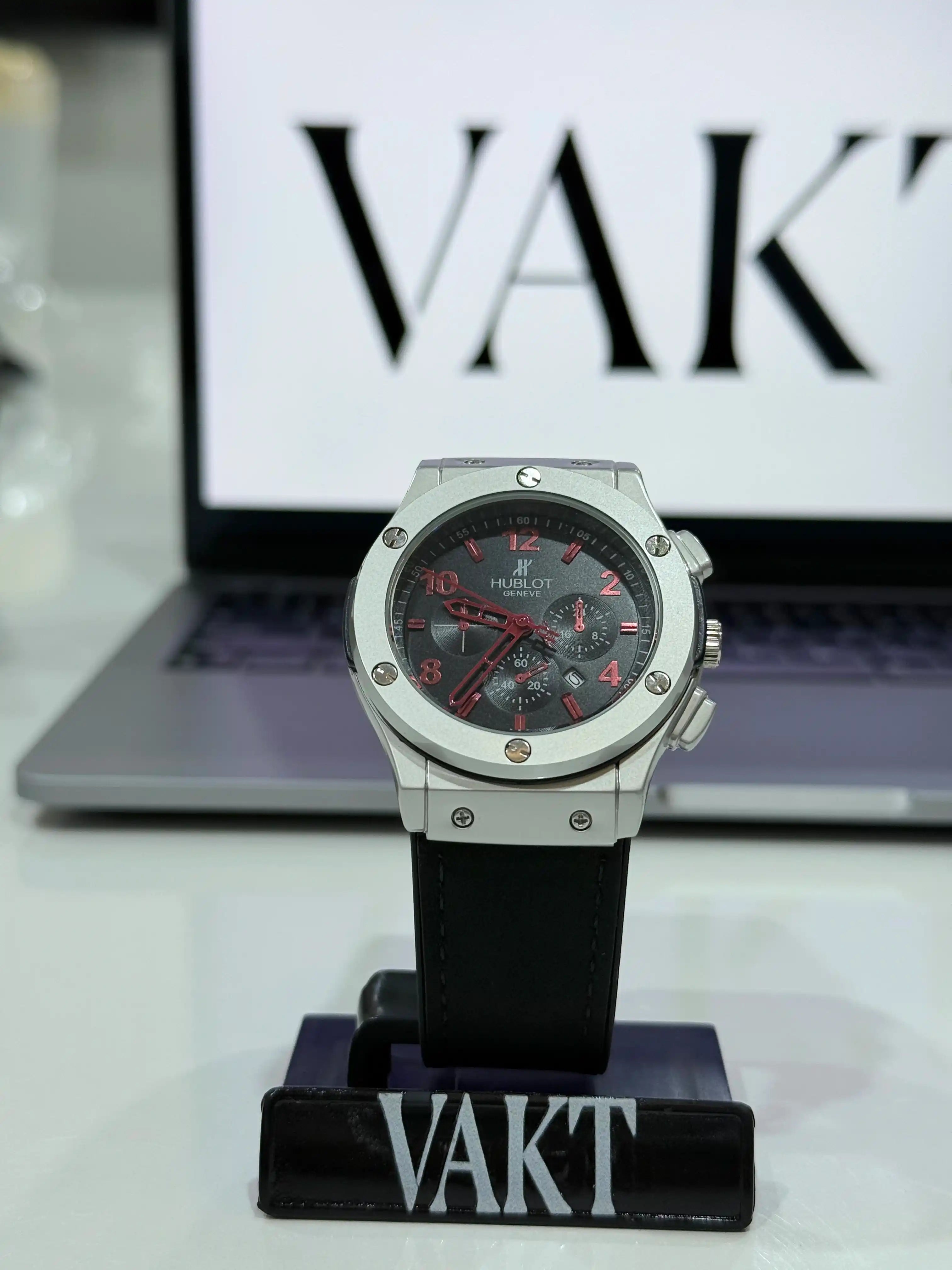 Silver Hublot Big Bang 44 mm Evolution watch with black strap on a stand, blurred 'VAKT' logo in the background