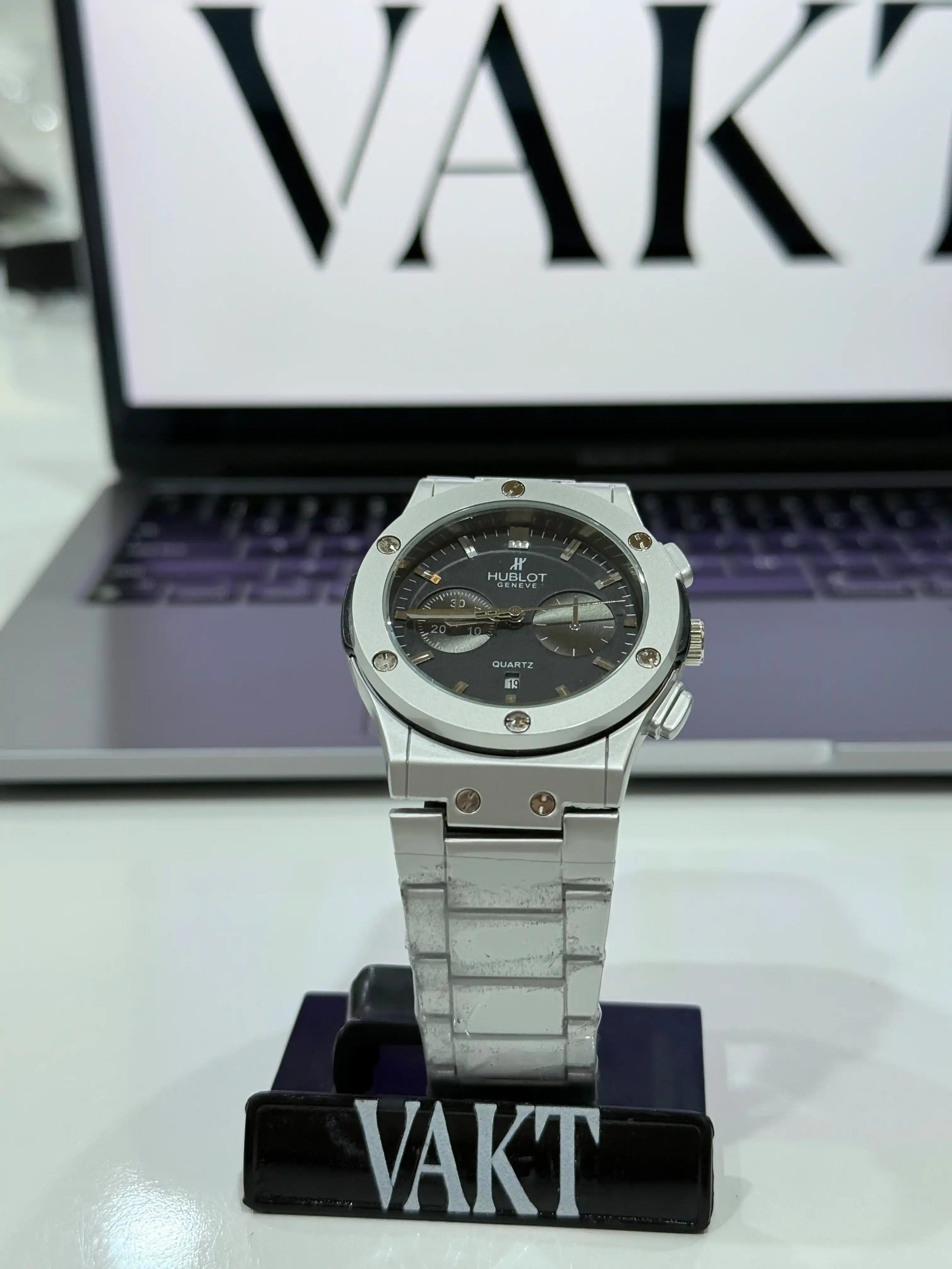 Silver hublot watch on a stand with a laptop displaying 'VAKT' brand logo in the background