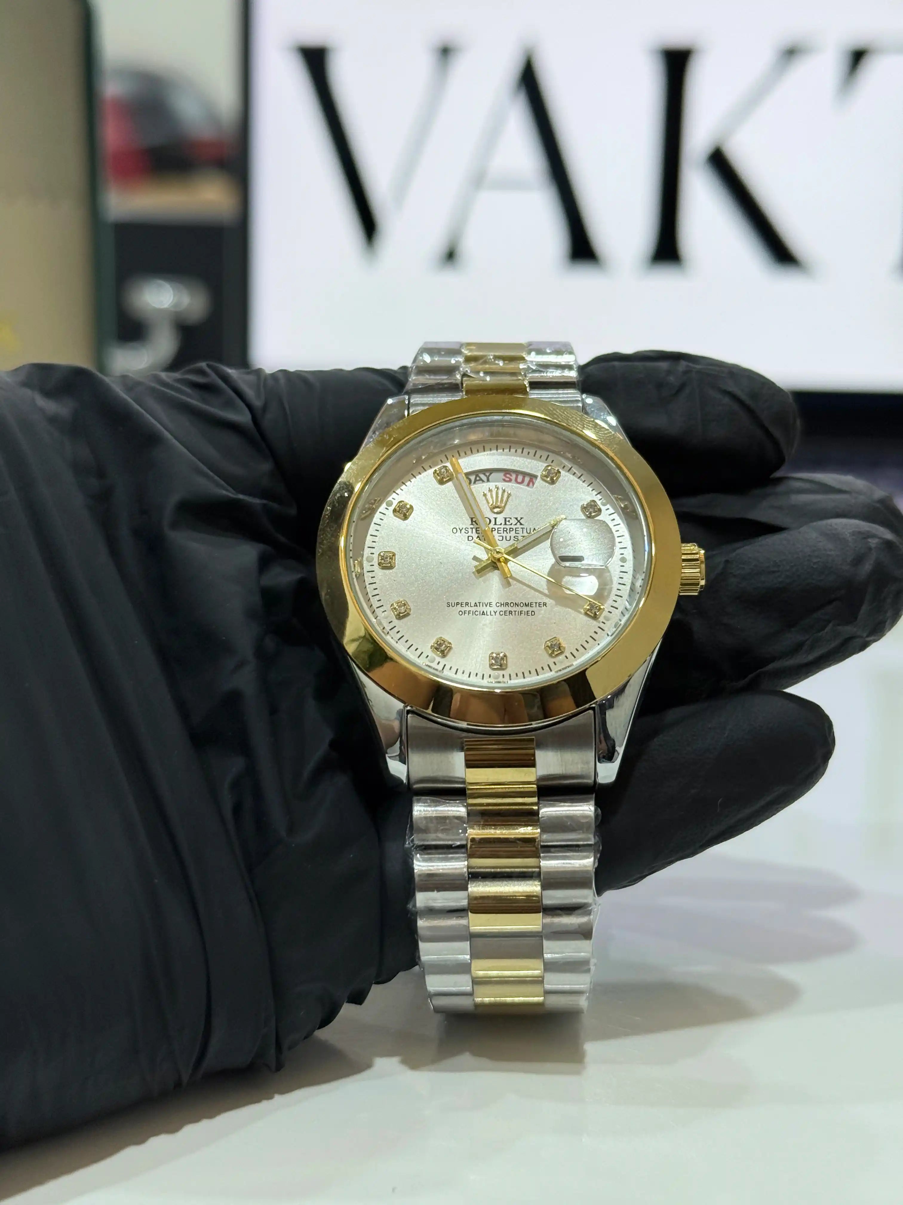 Gold and silver rolex perpectual day-date watch held by a gloved hand with 'VAK' logo in the background