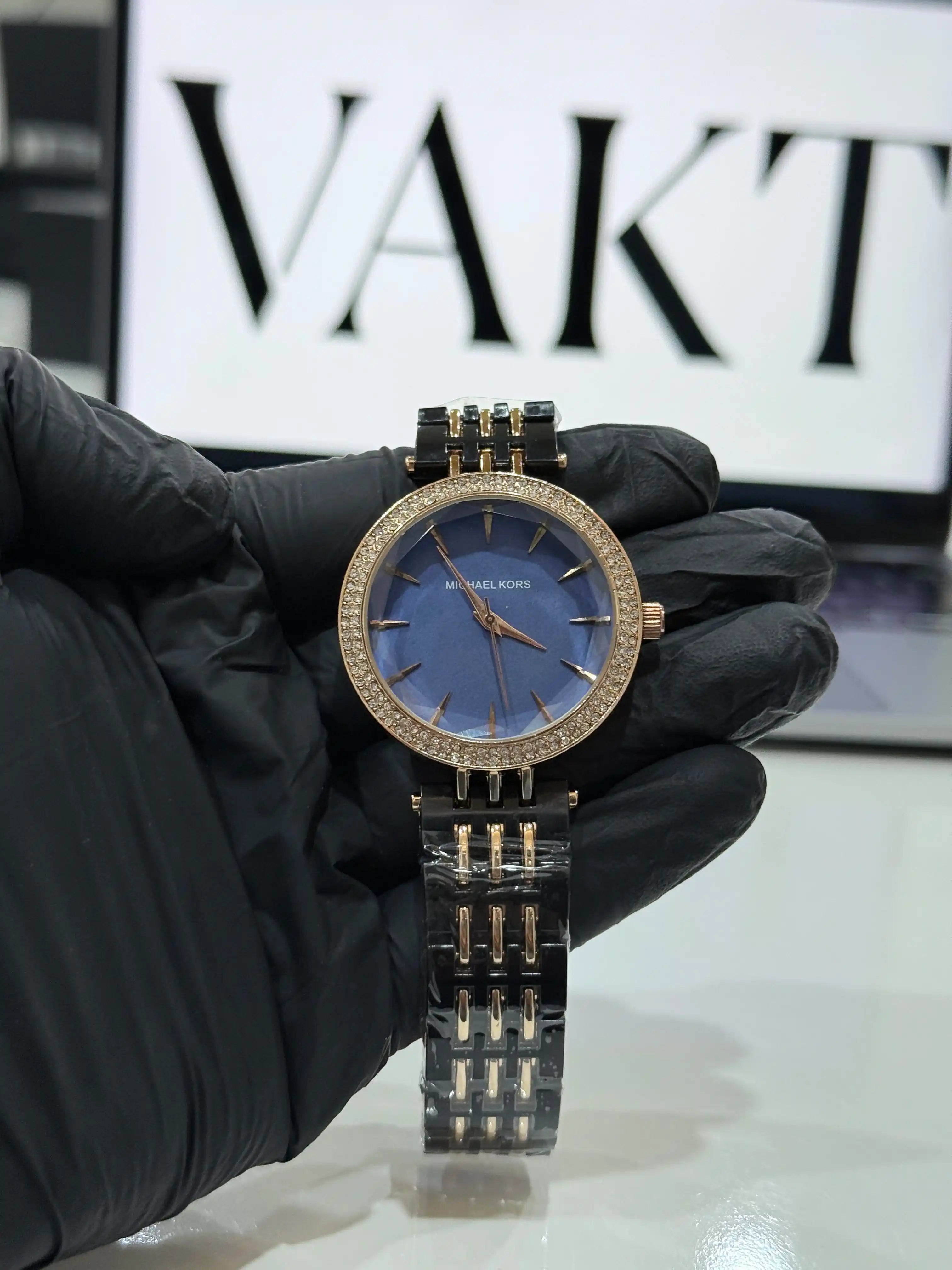Luxury michael kors darci watch with blue dial and gold accents held by a gloved hand, brand 'VAKT' visible in the background.