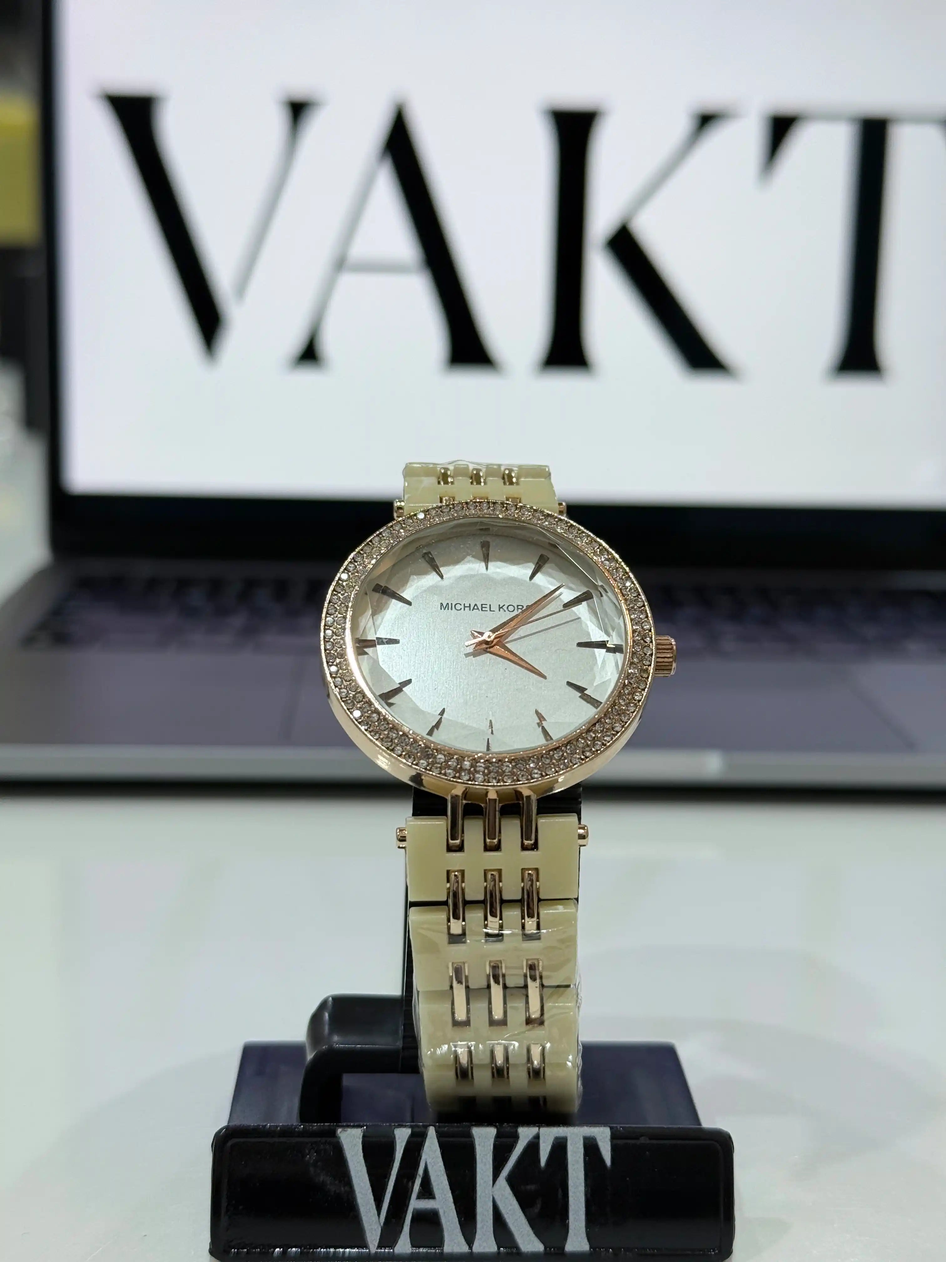 Michael Kors darci women's watch with a mother-of-pearl dial and a rose gold-tone stainless steel and pink acetate band on a stand with 'VAKT' branding in the background