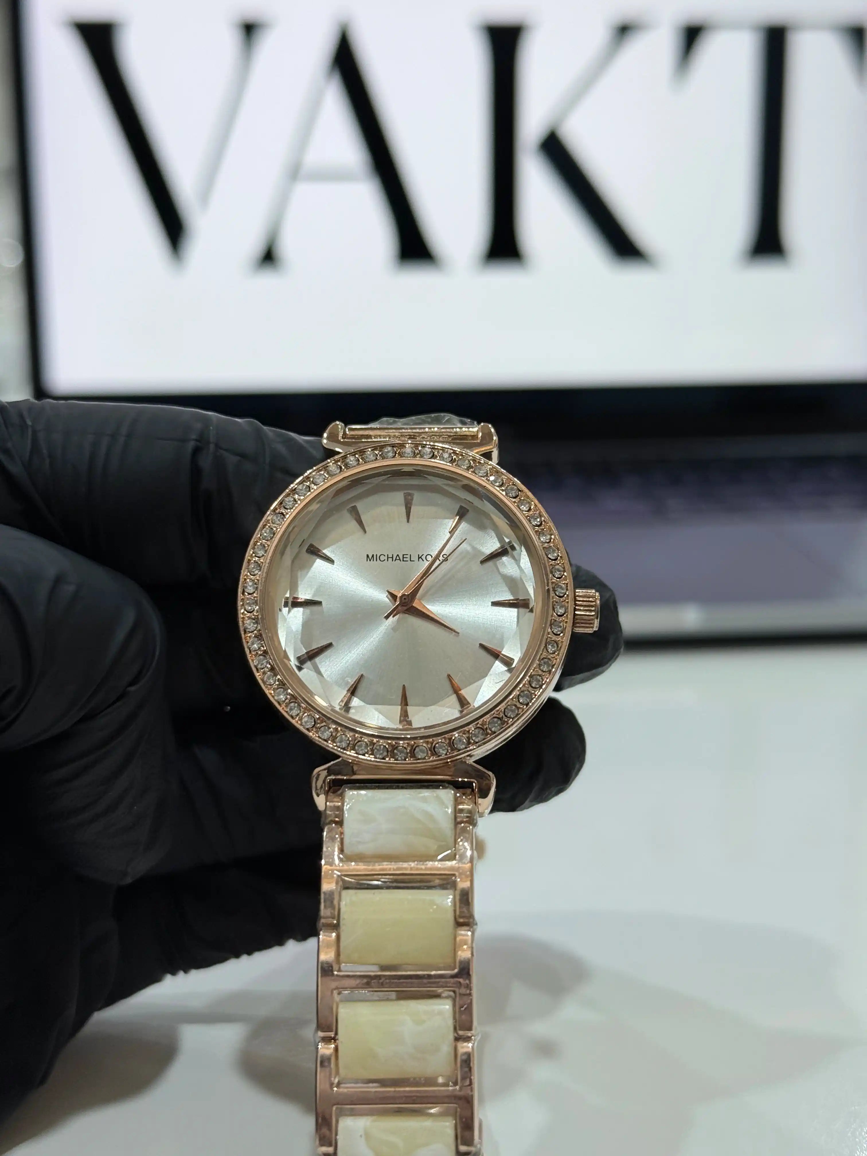 Rose gold MK watch with diamond accents held by a gloved hand, brand name 'VAKT' visible in the background.