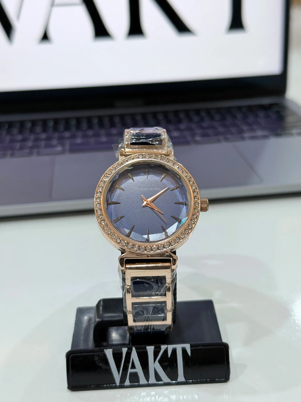 Rose gold watch with blue face on a stand in front of a laptop with 'VAKT' branding.
