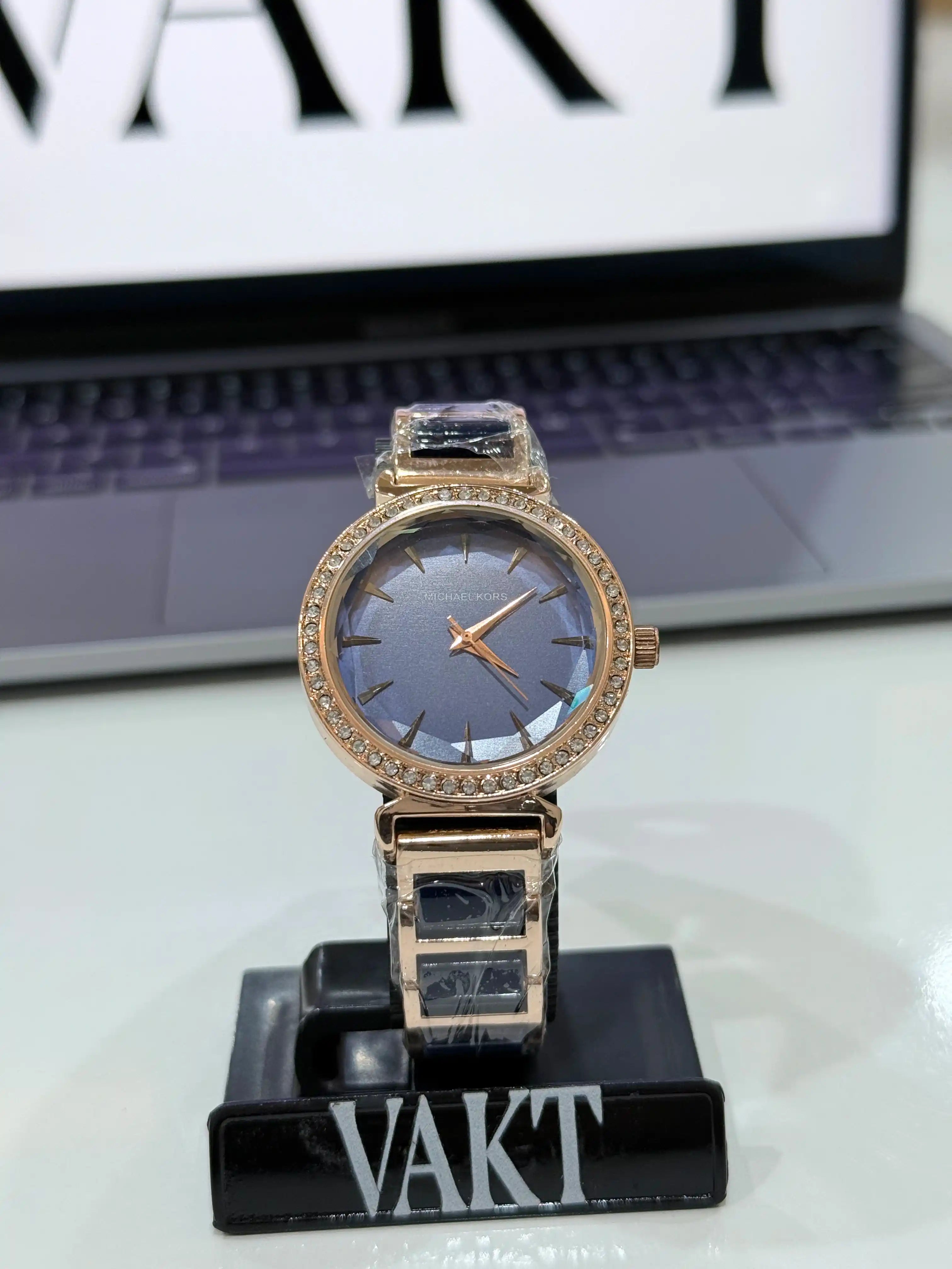 Rose gold watch with blue face on a stand in front of a laptop with 'VAKT' branding.