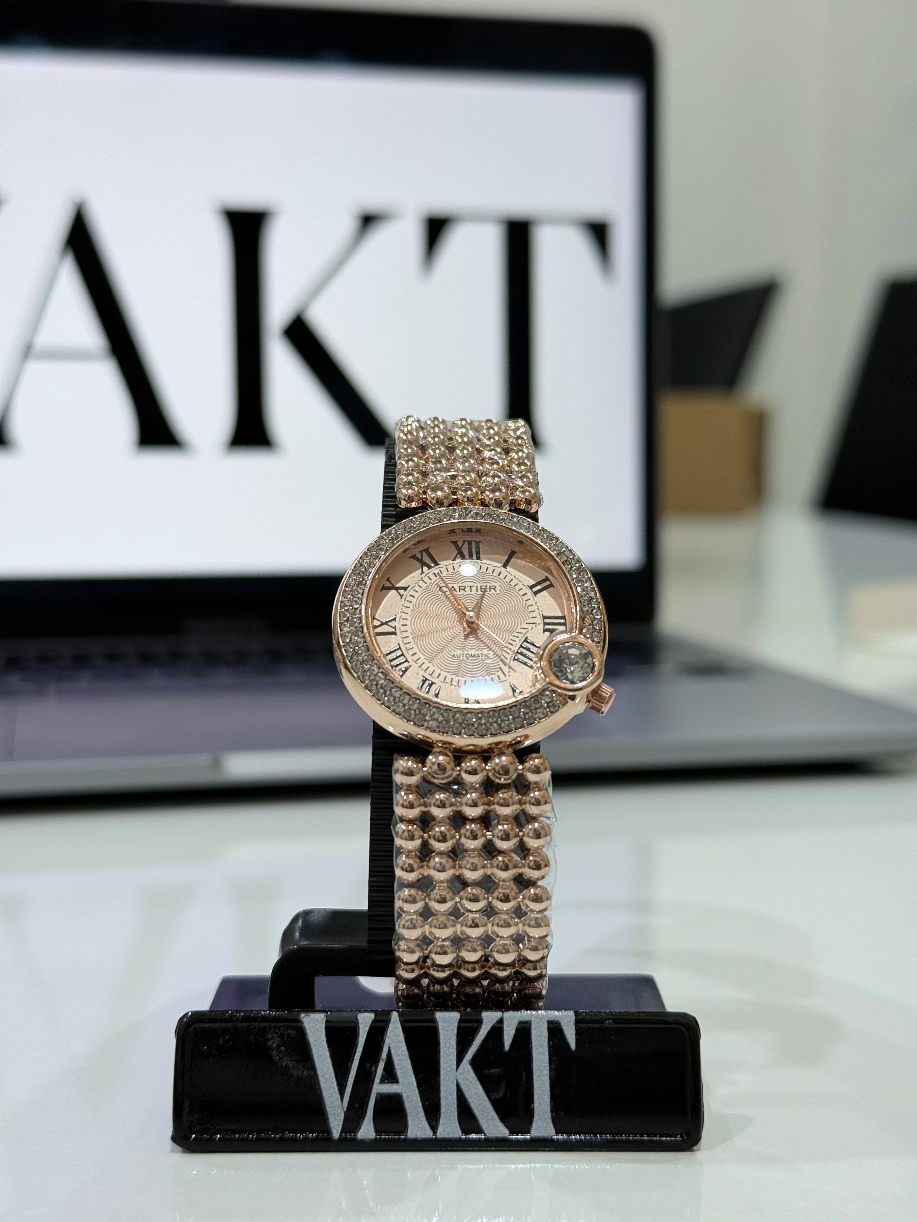 ladies wristwatch with gemstones on a stand in front of a laptop displaying 'VAKT'.