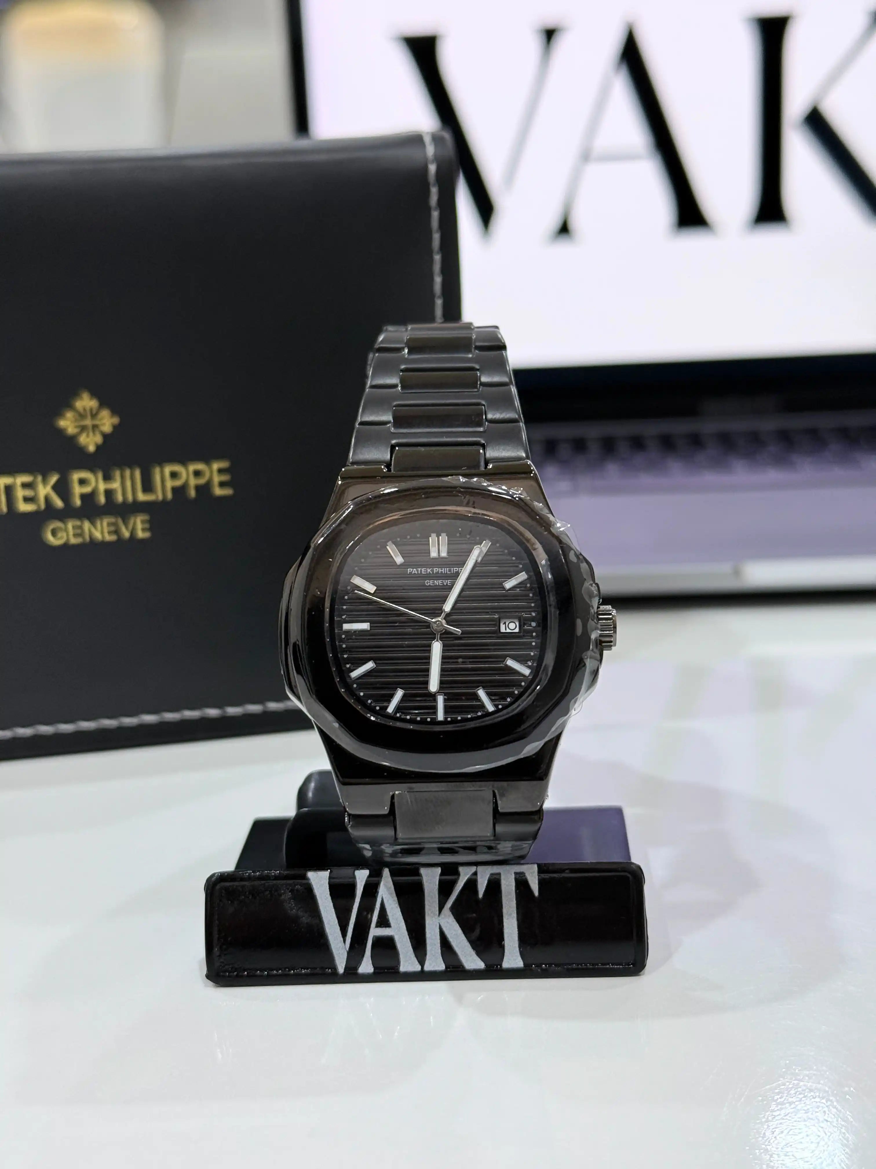 Black patek phillipe watch on a stand with 'VAKT' branding, against a blurred background with 'VAKT' logo.